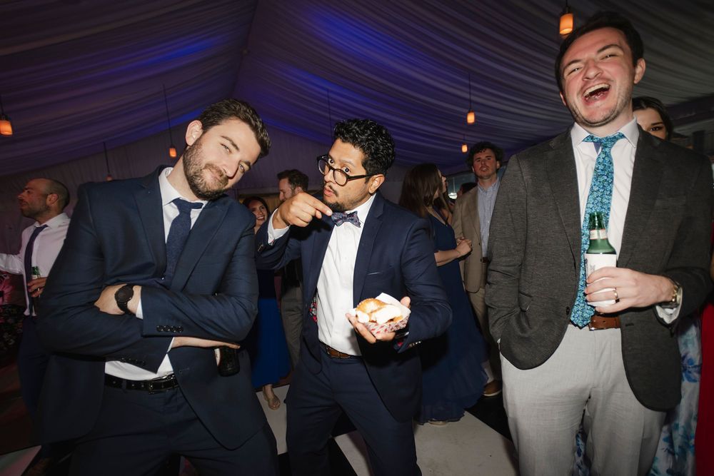 Three men in suits laugh and dance together at a formal wedding reception party.