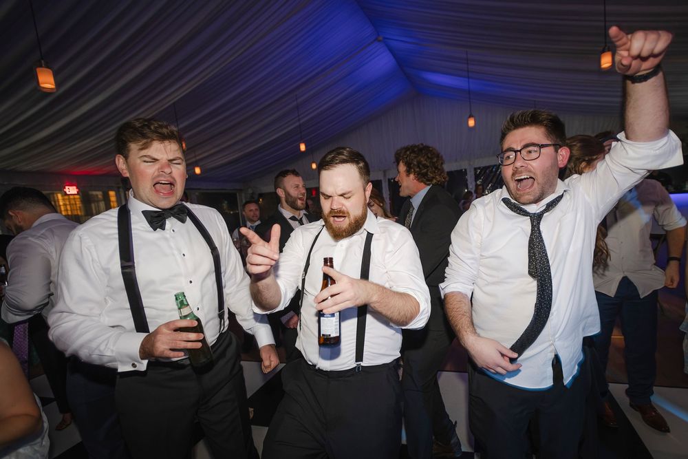 Three men in white shirts and suspenders dance and celebrate with beers at a wedding.