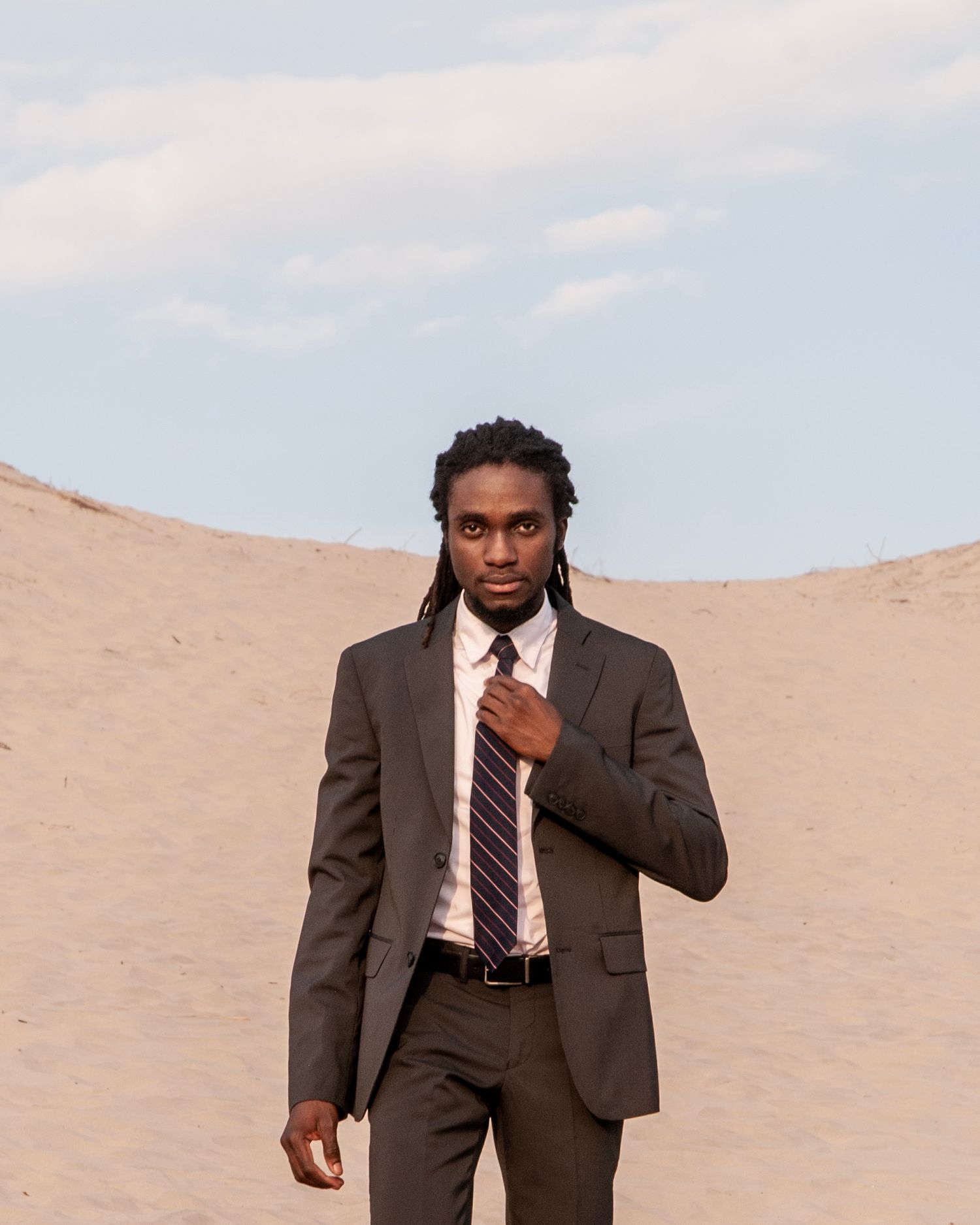 Stylish man in dark suit and striped tie stands confidently on sandy desert dunes under cloudy sky.