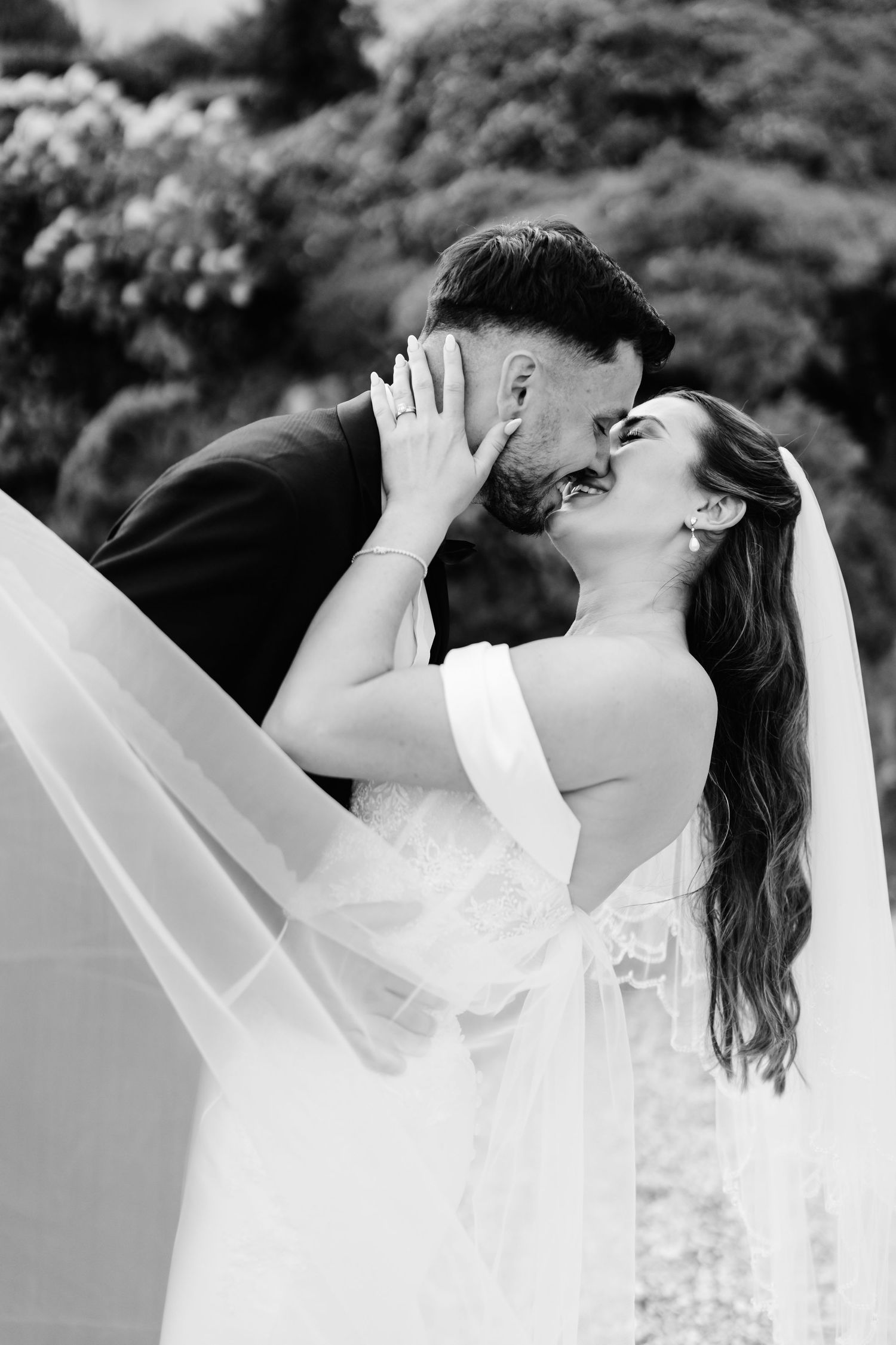Black and white photo of a bride and groom sharing a romantic kiss, with her veil flowing gracefully in the breeze.