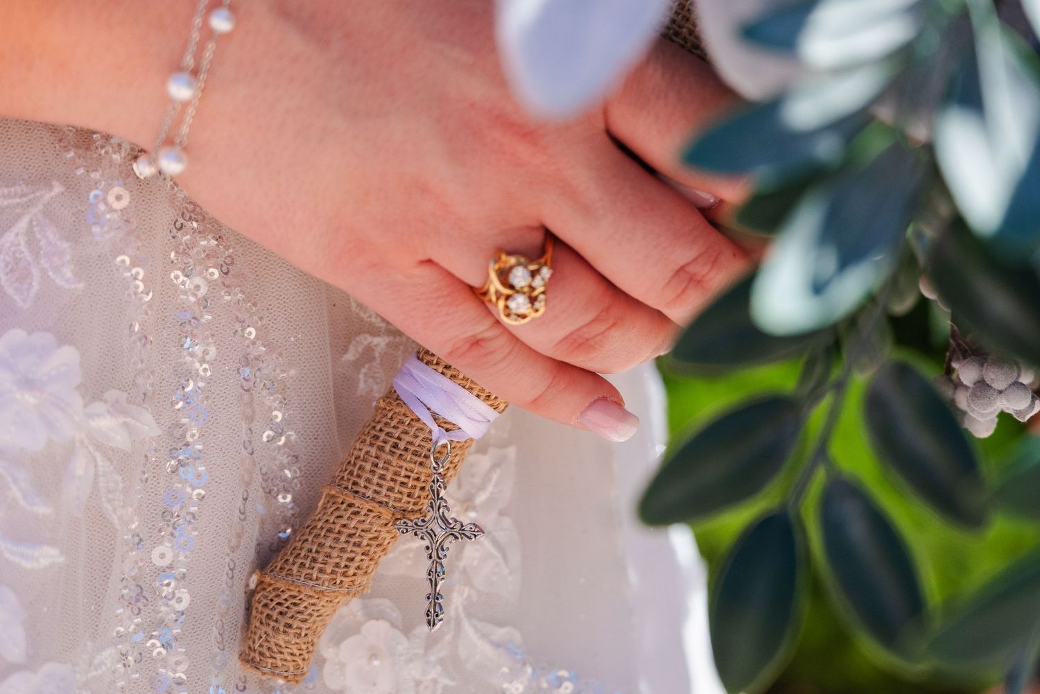 Close-up of a bride's hand wearing a gold floral ring, holding a burlap bouquet charm with a cross pendant.