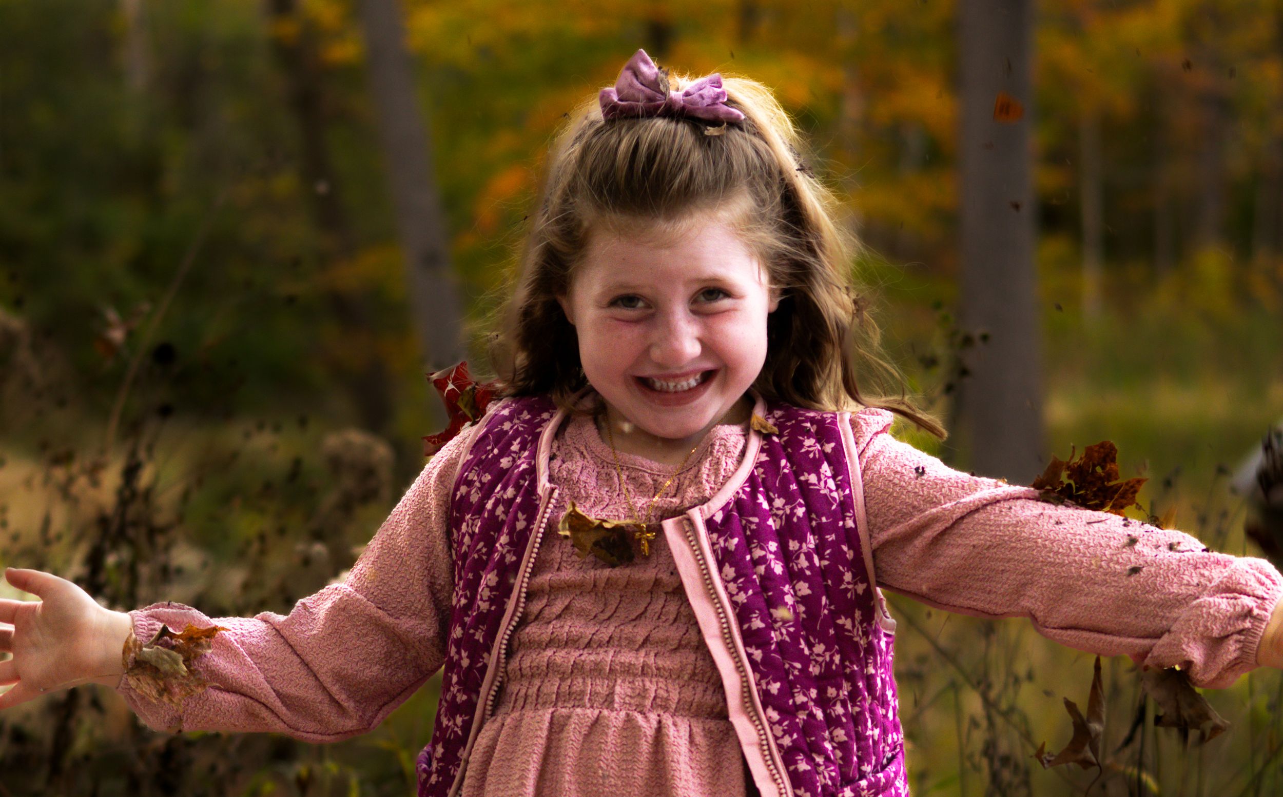 Happy young girl in pink outfit smiles with arms outstretched in autumn forest setting.