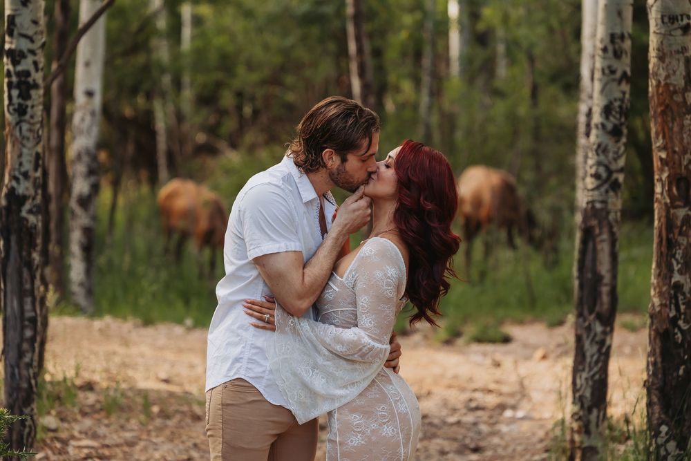 Couple sharing a romantic kiss in a forest clearing, woman with red hair in lace dress, horses grazing in background.