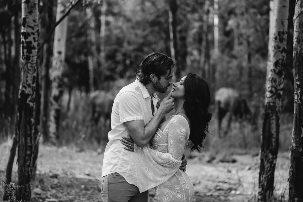 Black and white photo of a couple sharing a romantic kiss among birch trees in a forest setting.