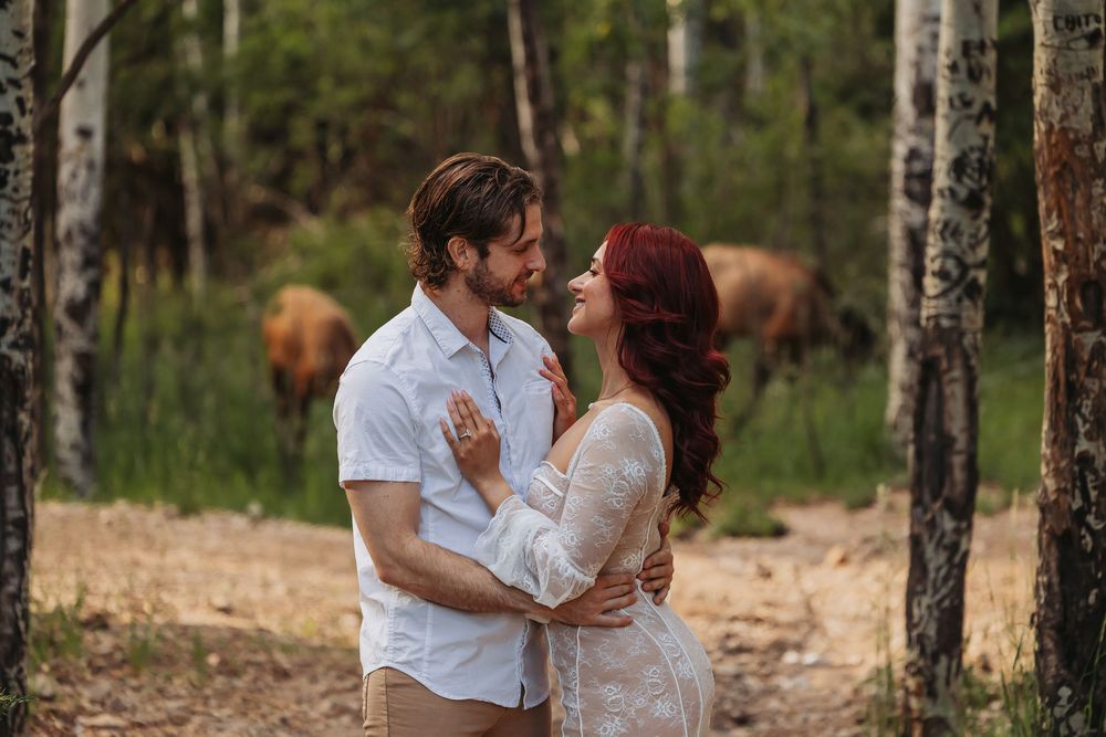 Couple embracing in a forest clearing, woman in lace dress with red hair, man in white shirt, cows grazing behind them.
