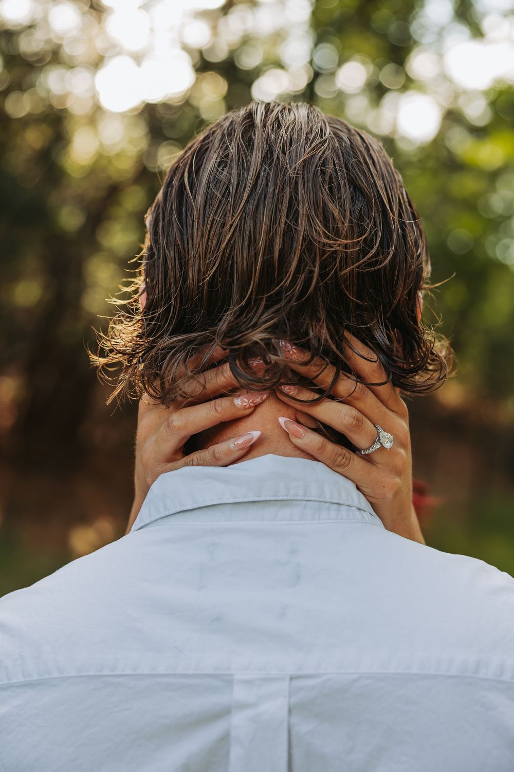 Back view of a person with brown hair, hands clasped behind their neck, wearing a white shirt outdoors.