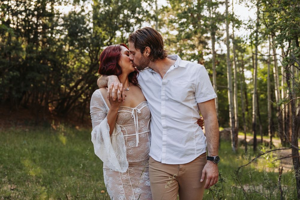 A couple shares a romantic kiss outdoors in a sunlit forest, the woman in a lace dress and the man in a white shirt.