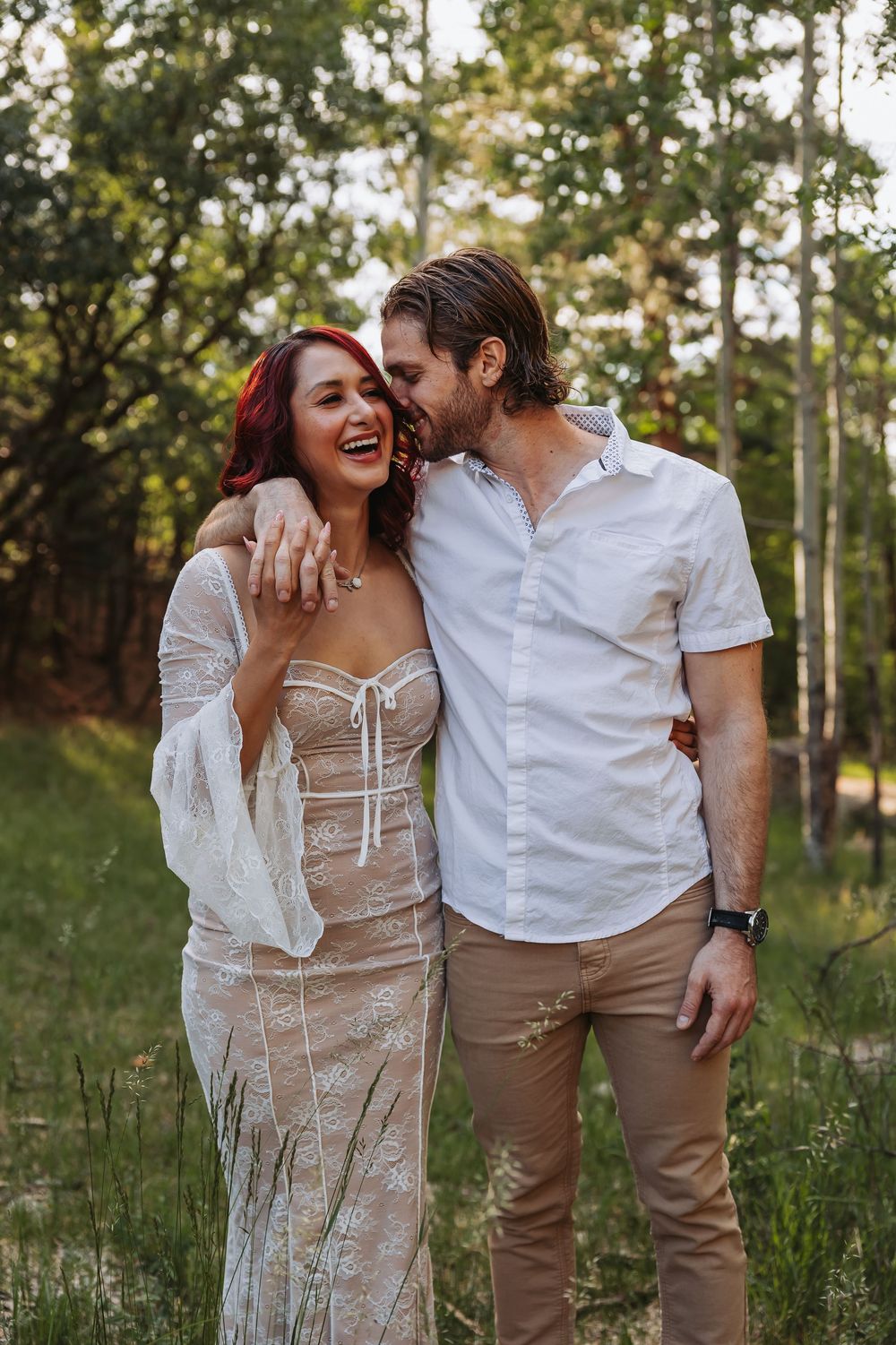 A laughing woman in lace dress and red hair embraces a smiling man in white shirt outdoors in a forest setting.