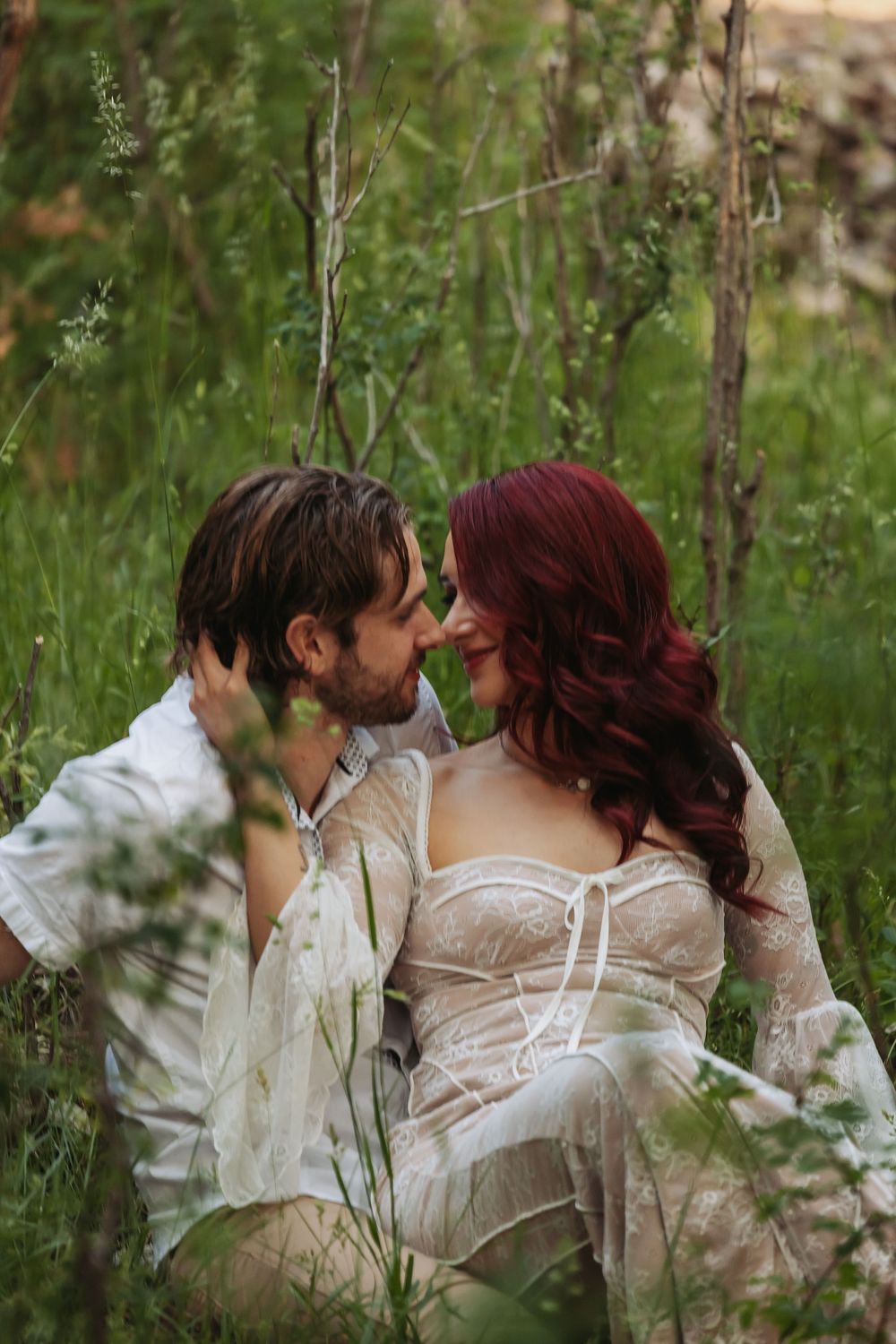 A couple sitting in lush green grass, leaning in for a kiss, woman with red hair in a white lace dress.