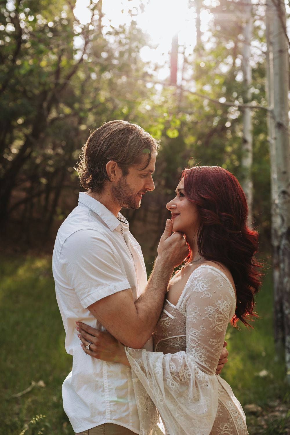 Couple gazes lovingly at each other in sunlit forest, man in white shirt touching womans chin, she wears lace dress.
