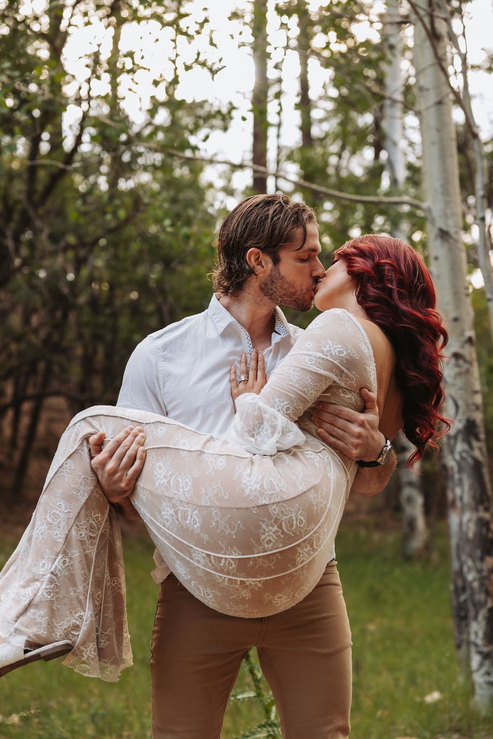 Man in white shirt carries woman in lace dress while kissing in a lush green forest setting.