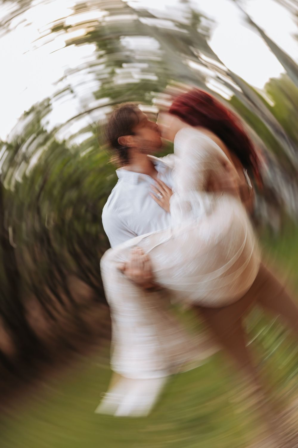 Blurred spin effect photo of a couple kissing and embracing outdoors, surrounded by green trees, creating a dreamy motion.