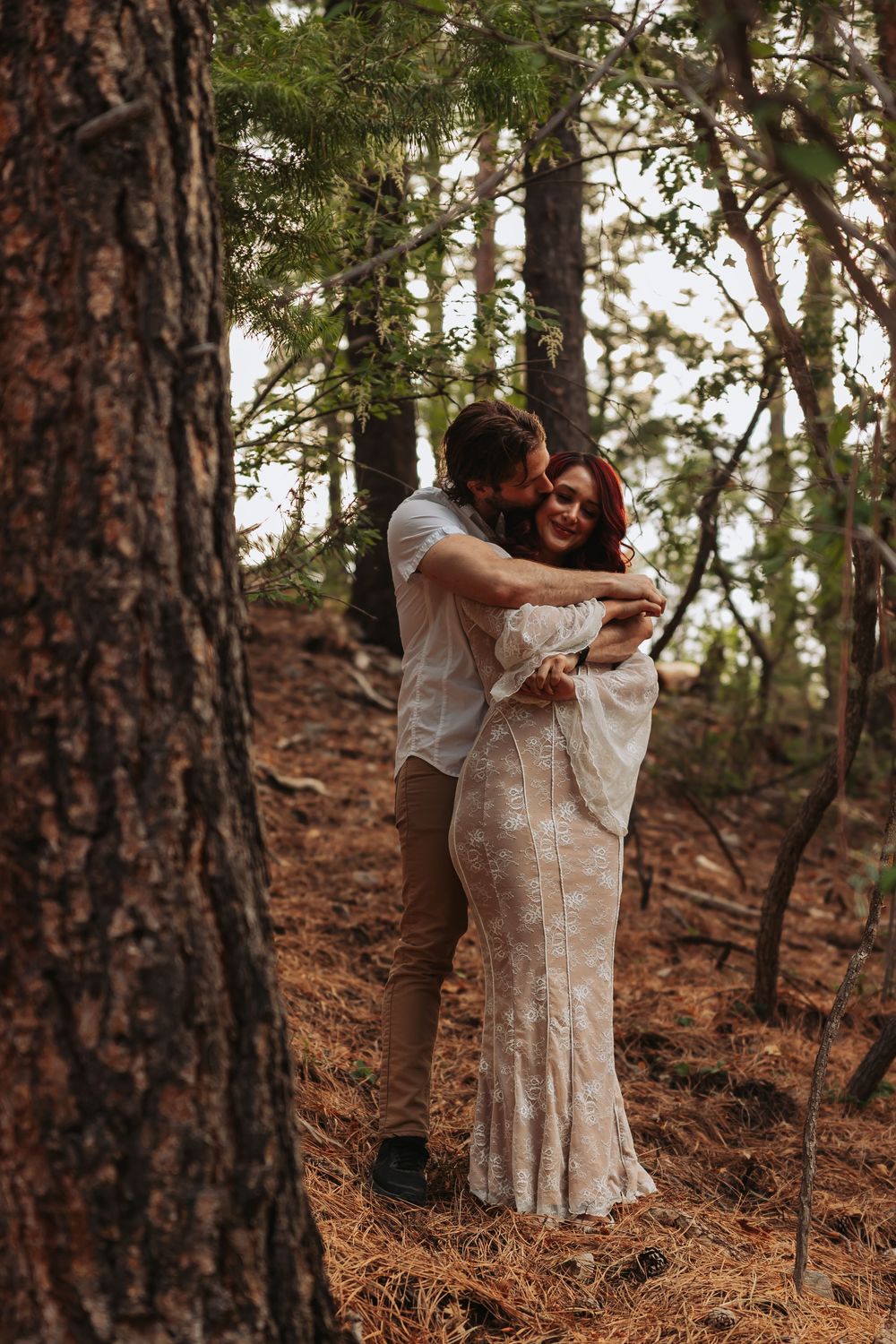 Couple embraces romantically among pine trees, man kissing womans head, she wears a boho lace gown.