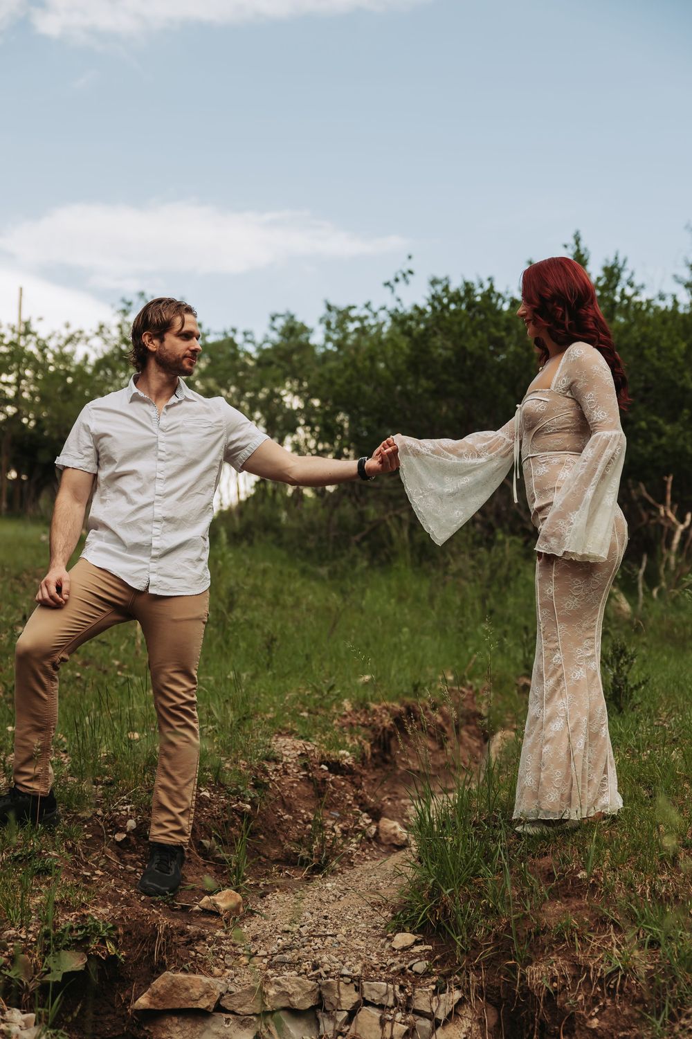 Couple holds hands on rocky path in green meadow, woman in lace dress with bell sleeves, man in khakis.