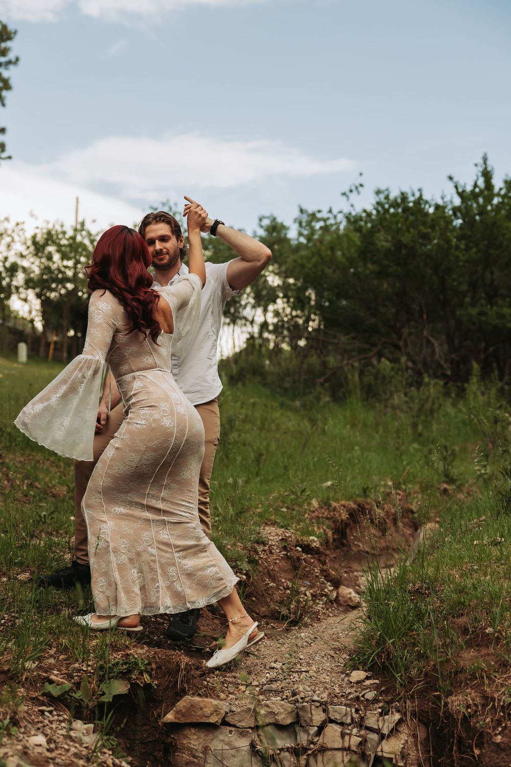A couple dances outdoors on a rocky path, woman in lace dress with red hair, man in white shirt, green trees behind.