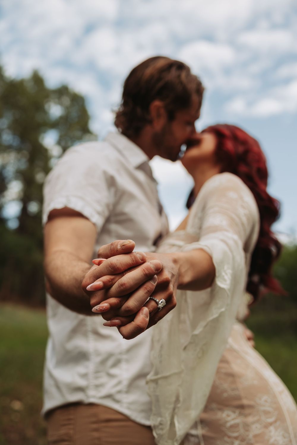Couple kissing outdoors while holding hands showing engagement ring, red-haired bride in white dress with lush green background.