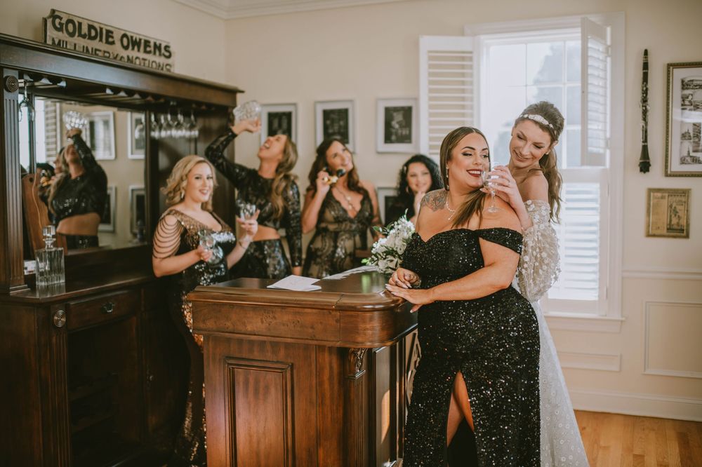 Bride and bridesmaids in black sequin gowns celebrating at a vintage wooden bar, raising glasses in a elegant venue.