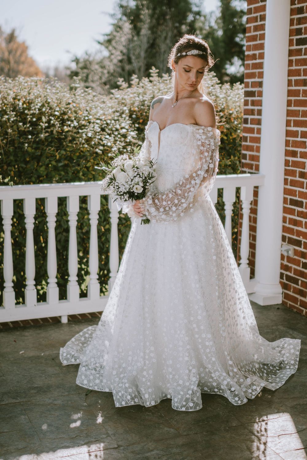 Bride in off-shoulder white lace ball gown with floral sleeves holds bouquet on a sunlit porch.