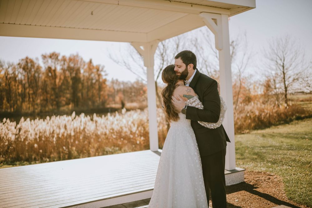 A newlywed couple shares a romantic kiss under a white gazebo surrounded by golden autumn foliage near a serene pond.