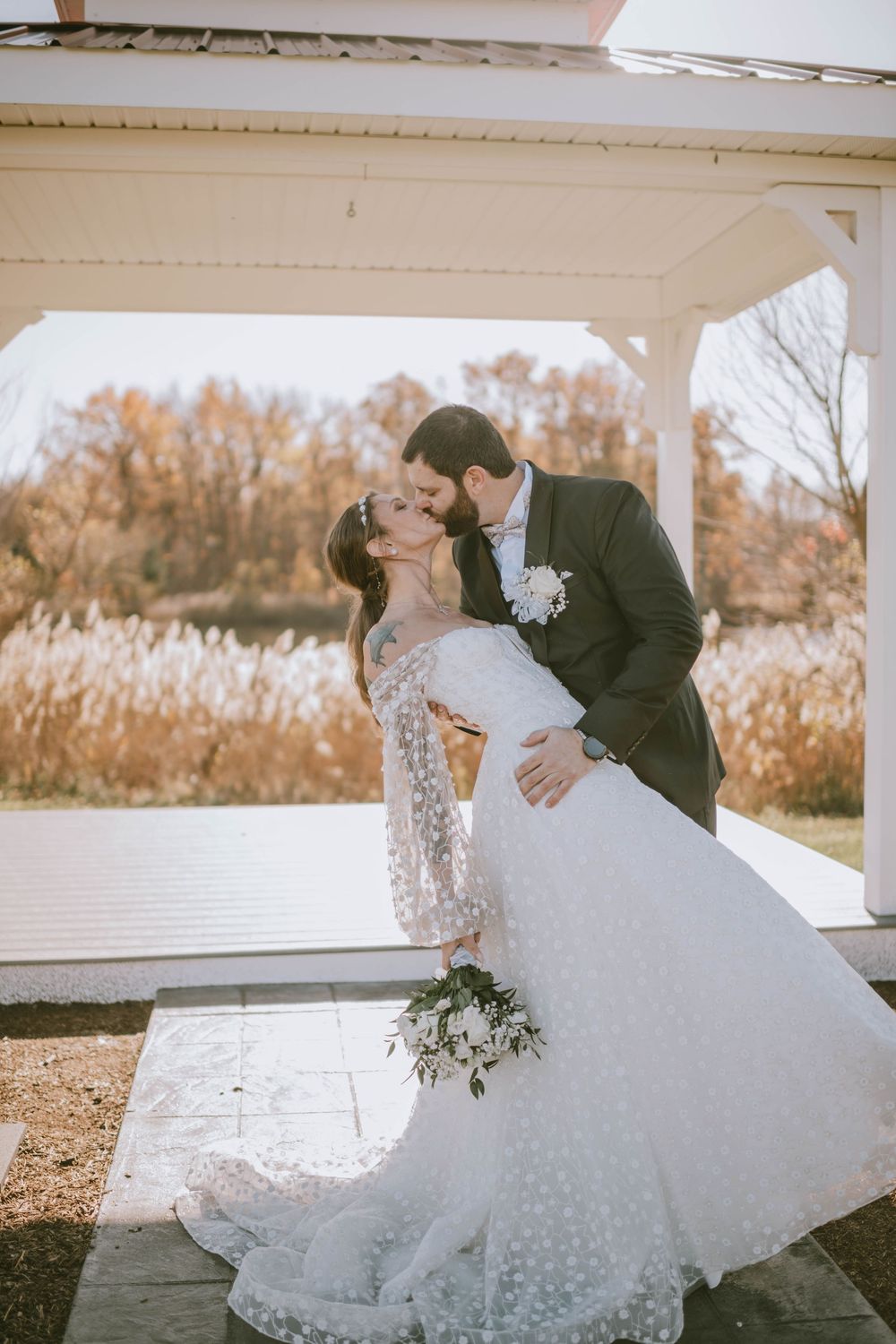Bride and groom share a romantic kiss under a white gazebo, surrounded by autumn foliage, as bride holds a white bouquet.