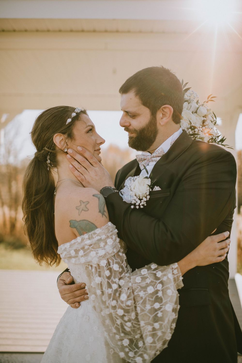 A bride and groom share a romantic embrace in golden sunset light, with floral boutonniere and lace wedding dress.