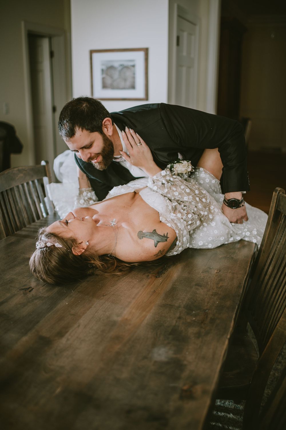 A groom leans over his bride lying on a wooden dining table, both smiling in wedding attire during a playful moment.