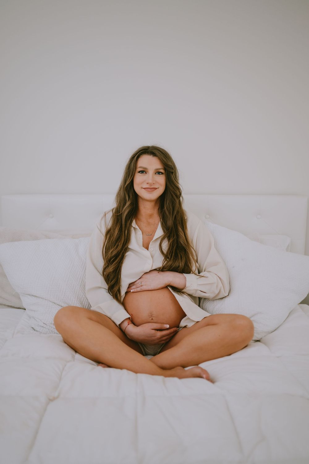 Pregnant woman sitting cross-legged on white bed, cradling bare baby bump, wearing open cream shirt, long brown hair.