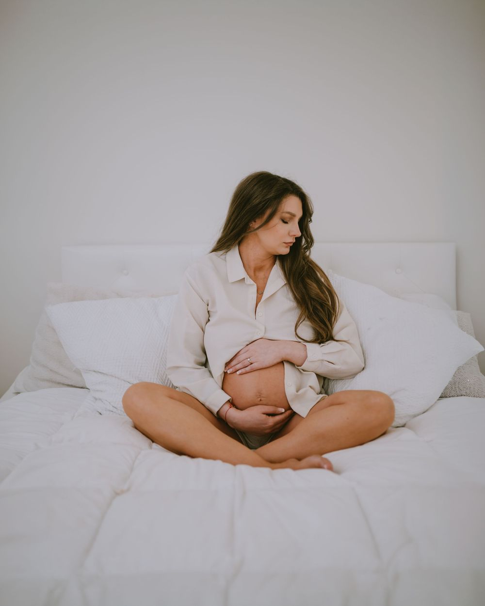 Pregnant woman sitting cross-legged on white bed, cradling baby bump in white shirt, serene maternity photo.