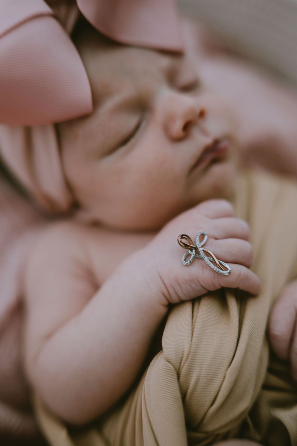 Sleeping newborn baby wearing a pink bow headband with a delicate cross ring on tiny fingers, wrapped in a soft golden blanket.