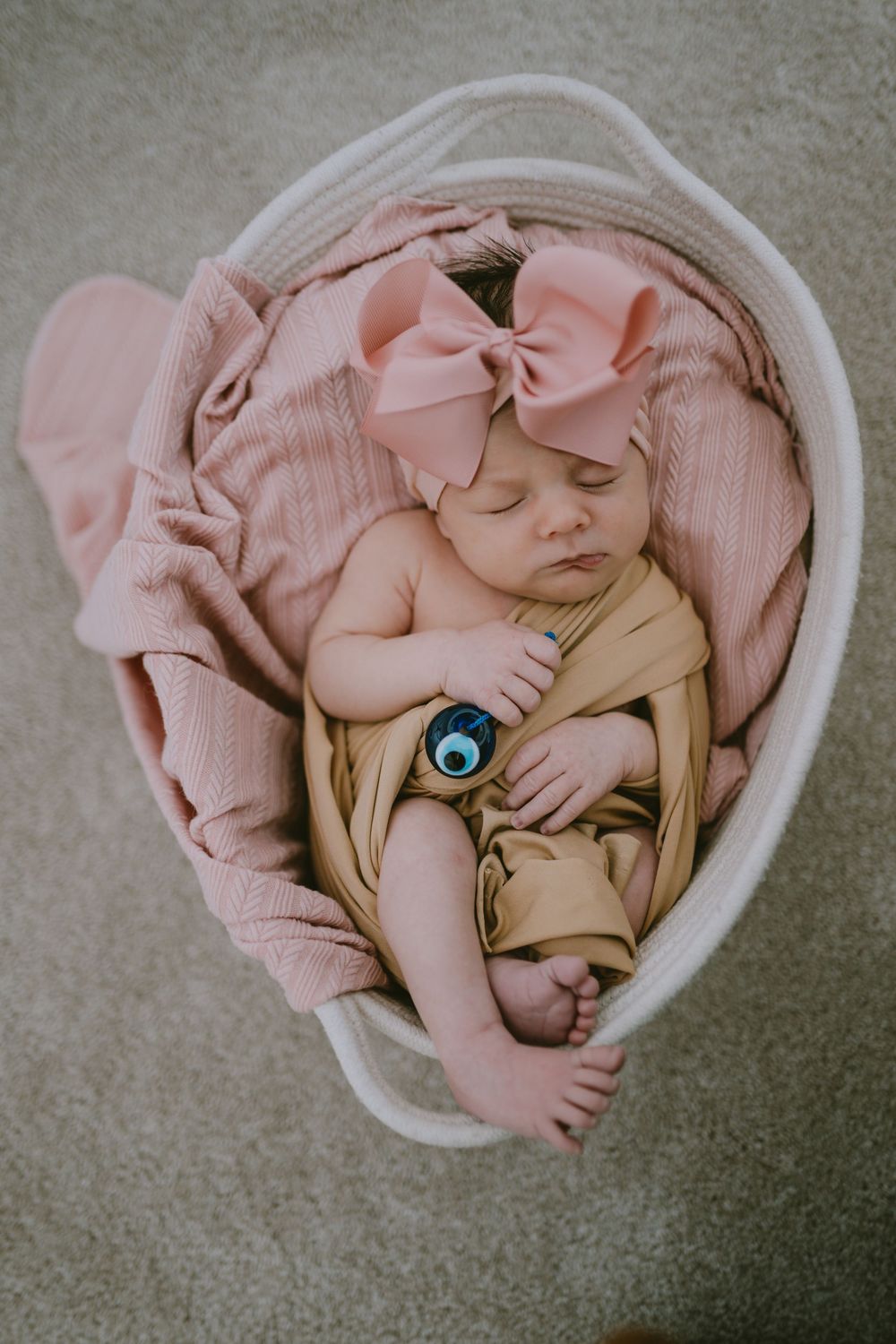 Newborn baby girl sleeping in a white basket with pink blankets and a large pink bow headband.