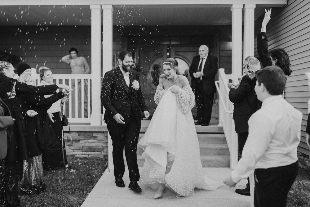Black and white photo of a bride and groom exiting a house as guests celebrate with confetti at a wedding.