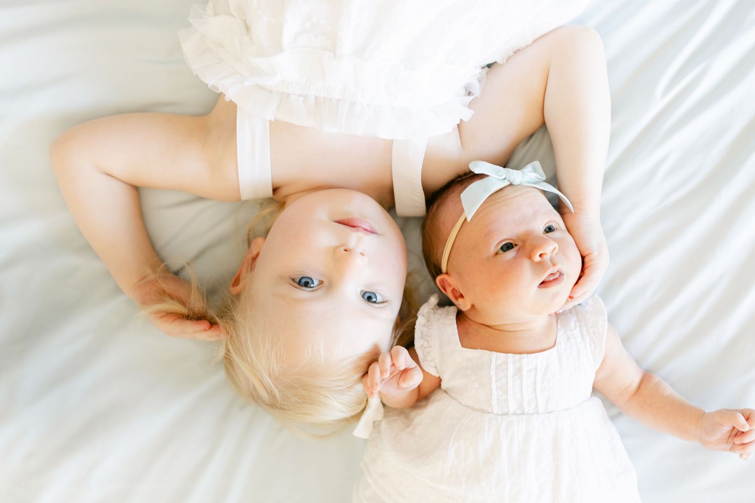 Two adorable baby girls in white dresses lying on a white bed, smiling and looking up at the camera.