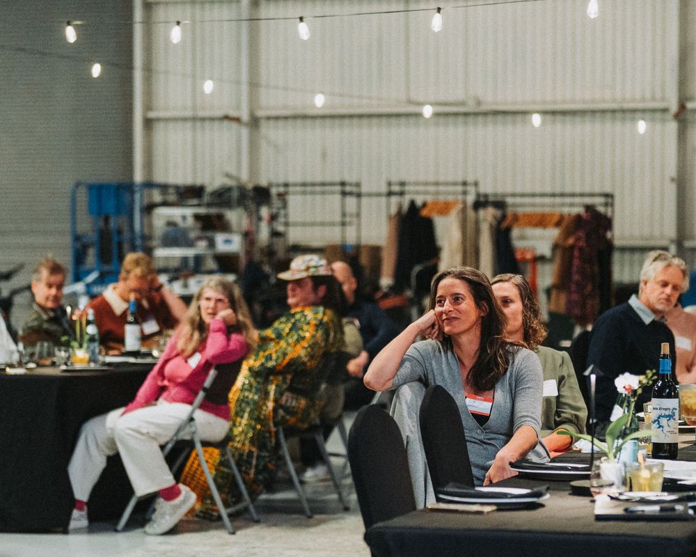 People seated at tables in an industrial warehouse venue during a dining event with string lights overhead.
