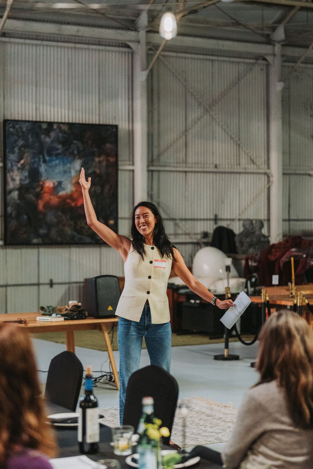 Enthusiastic woman presenter raising her hand while speaking at a Good People Dining event in a warehouse.