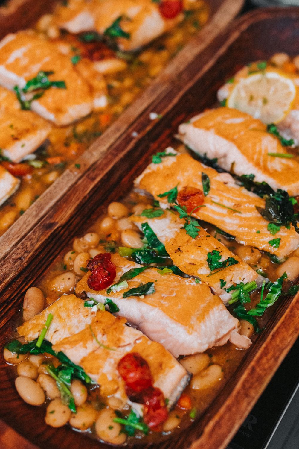 Baked salmon fillets with white beans, cherry tomatoes, and fresh herbs served on wooden platters.