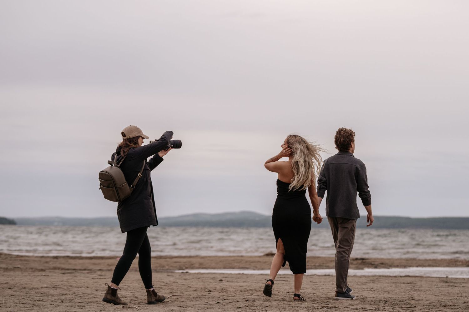 Photographer capturing couple walking on sandy beach near ocean under overcast sky.