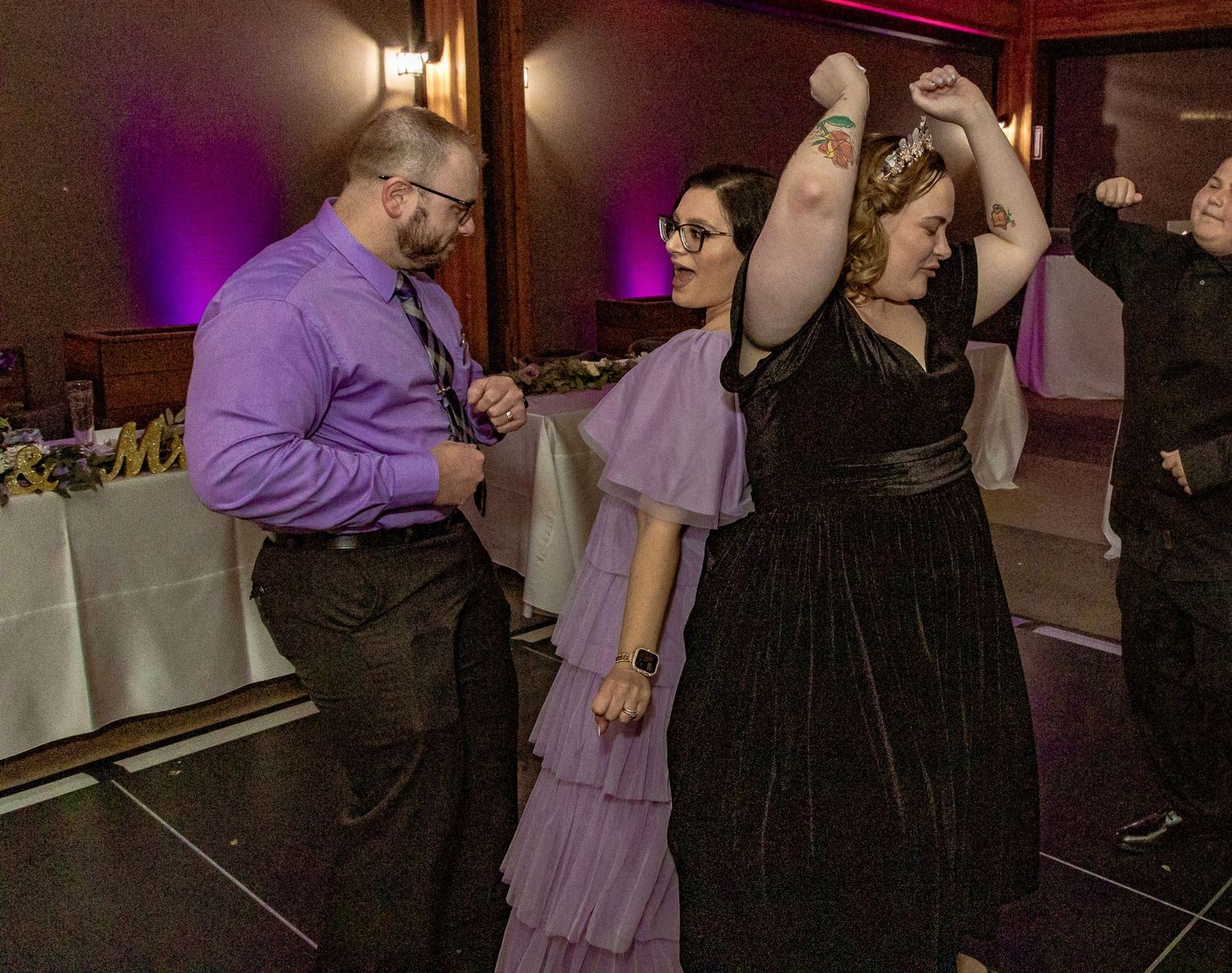 People dancing in formal attire at an indoor event, wearing purple and black dresses with colorful ambient lighting.