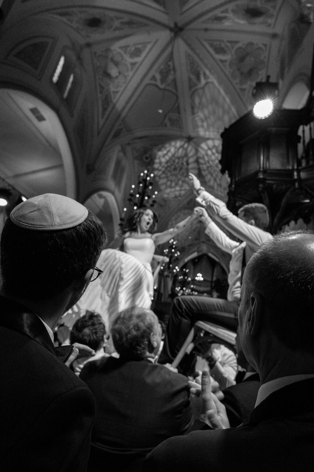 Black and white photo of a joyful Jewish wedding celebration with guests lifting the bride and groom in a grand vaulted hall.