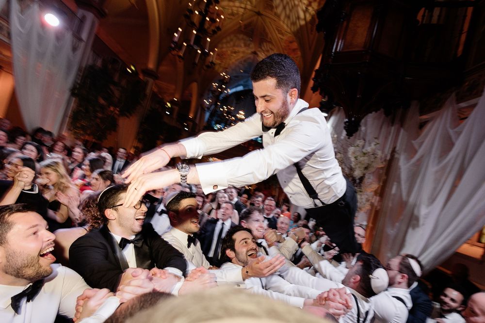 Groom crowd surfing at elegant wedding reception surrounded by cheering guests in formal attire.