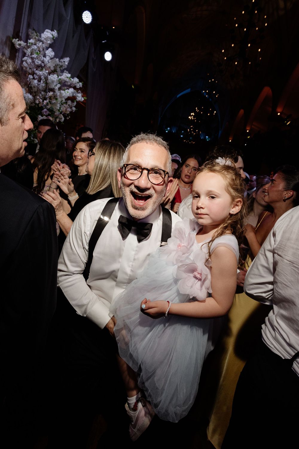 A joyful man in a bow tie and suspenders holds a young girl in a pink tutu at a festive celebration.