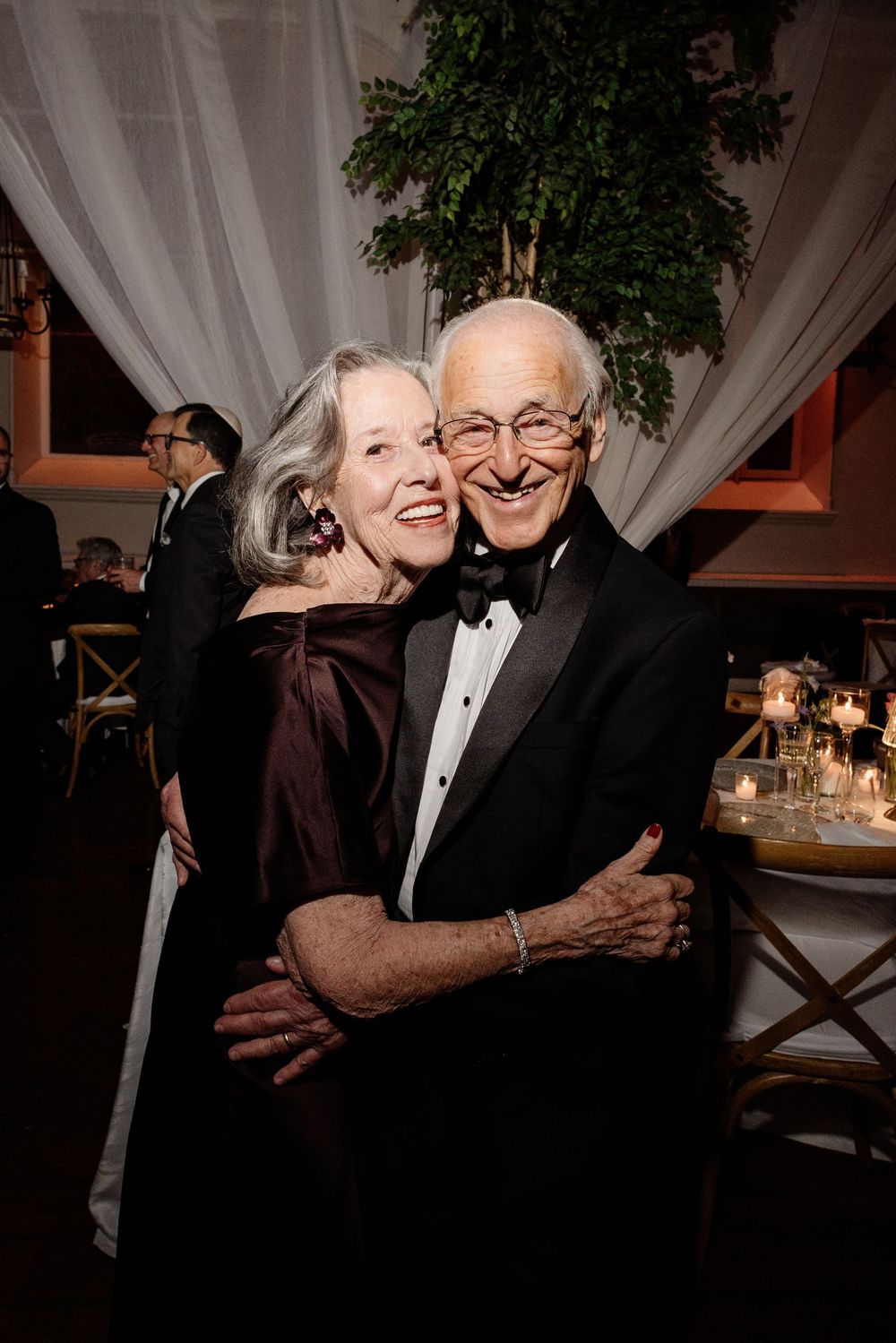 Elegant older couple embracing and smiling at formal wedding reception in black tie attire.
