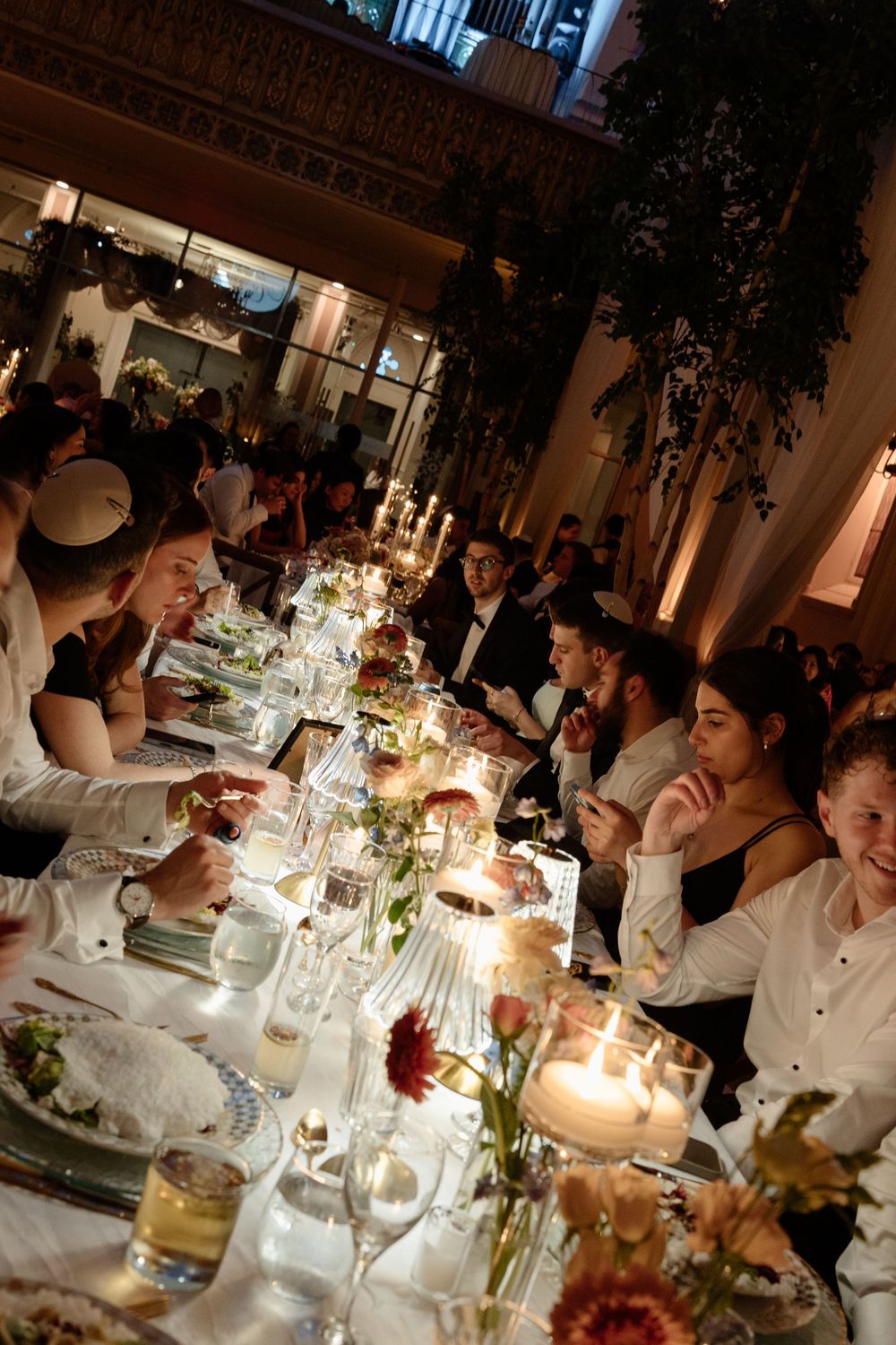 Elegant Jewish Seder dinner party with guests dining by candlelight at a long decorated table with flowers.