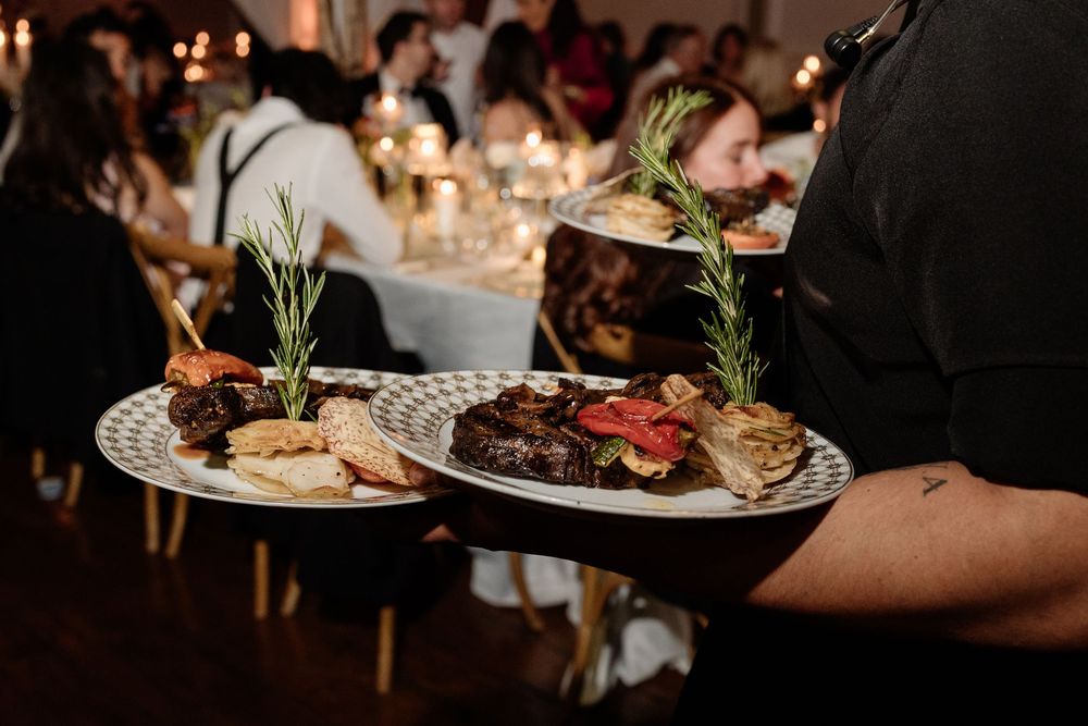 Waiter carrying elegant plates of gourmet meat dishes garnished with rosemary at a candlelit wedding reception.
