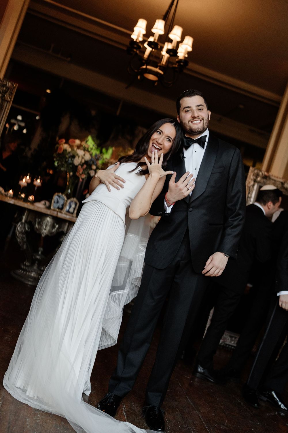 Bride in white gown and groom in black tuxedo smiling and showing wedding rings at elegant reception.