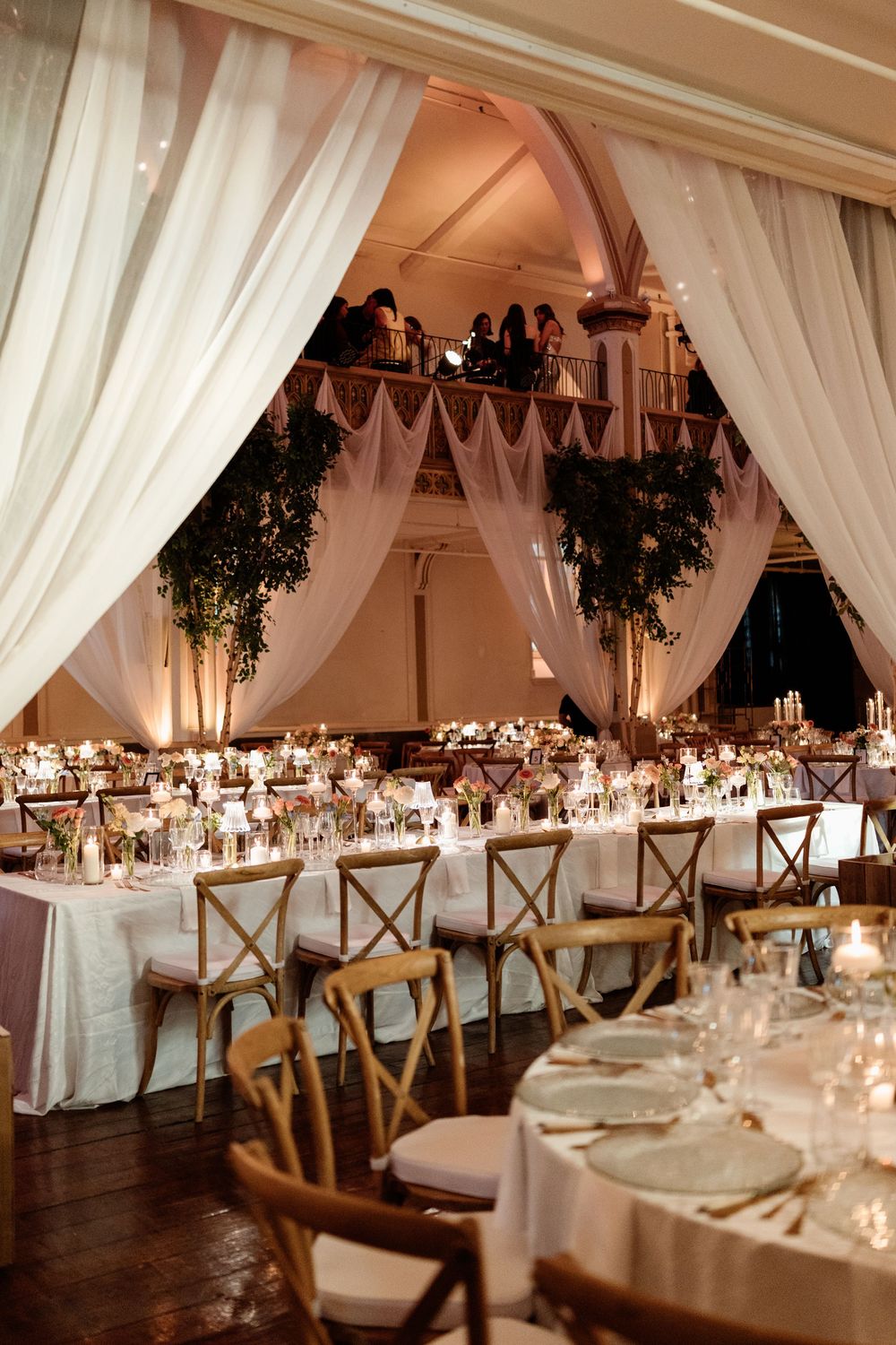 Elegant wedding reception hall with white draping, wooden cross-back chairs, candlelit tables, and lush greenery accents.