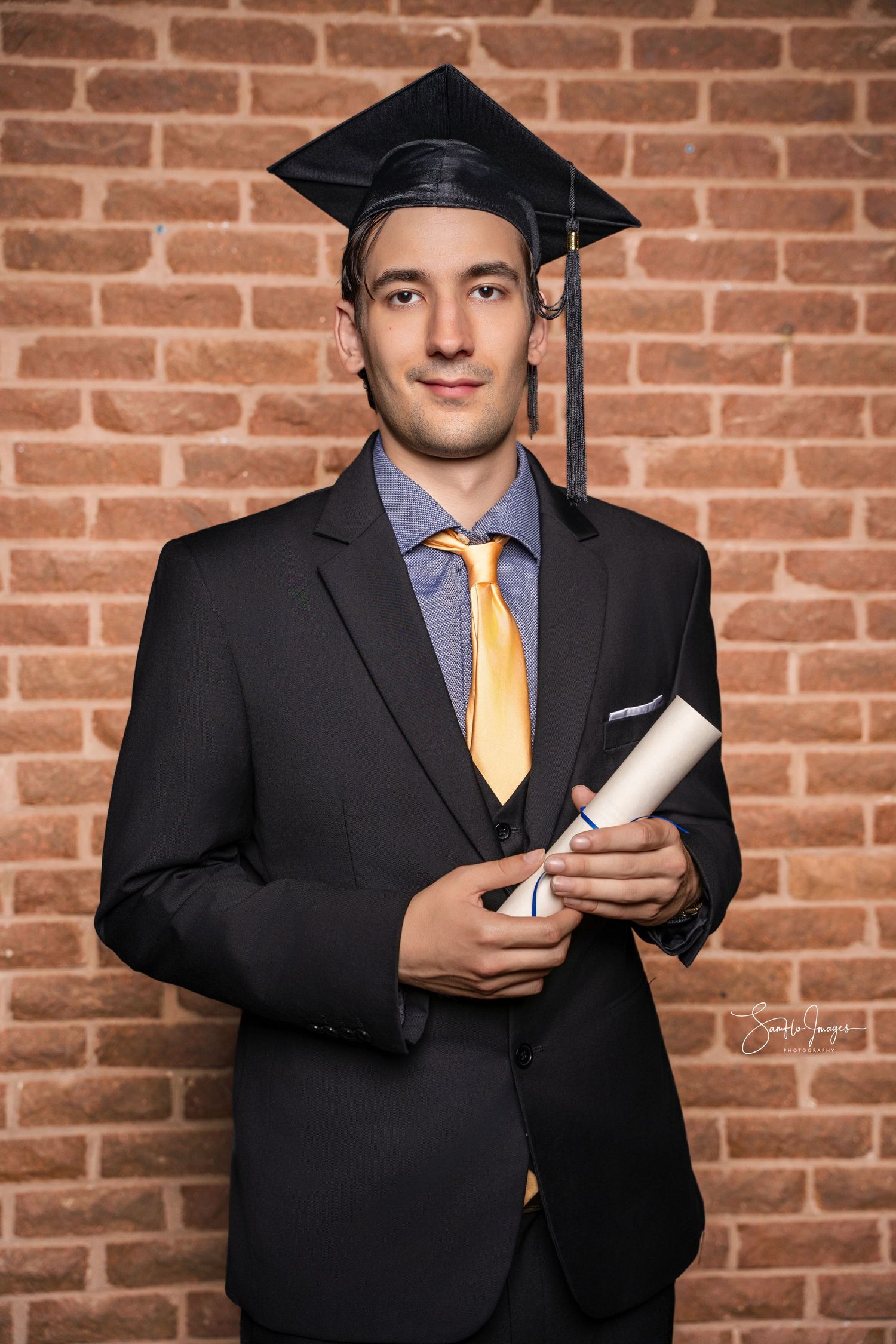 Young man in black graduation cap and gown holding diploma scroll, posing in front of brick wall.