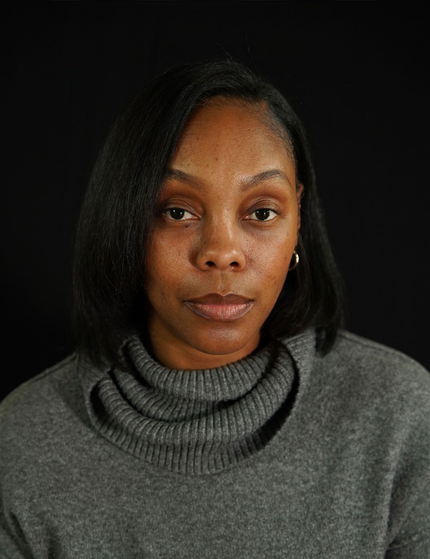 A woman with a black bob hairstyle wearing a gray turtleneck sweater poses against a dark background.