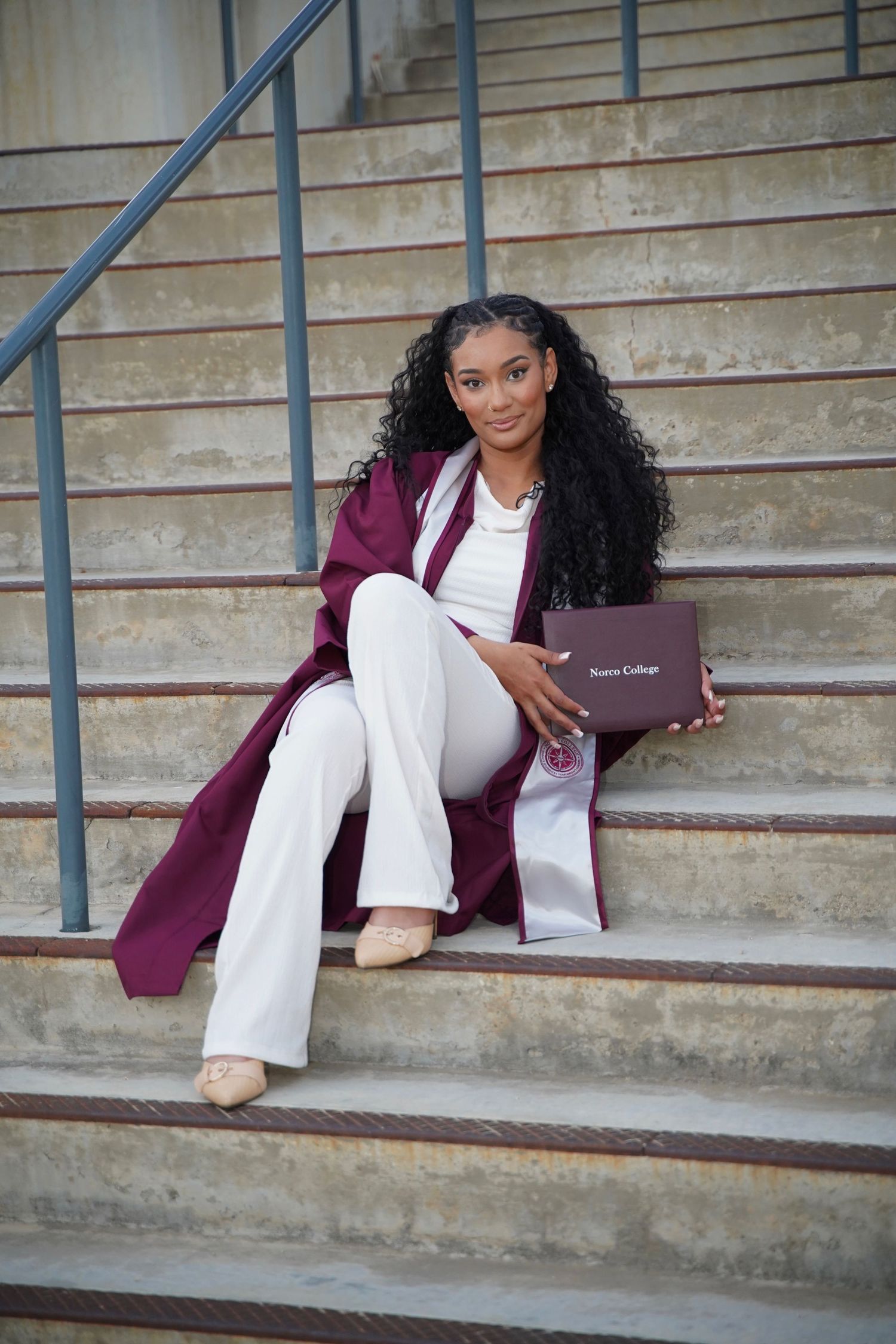 Young woman in maroon graduation gown sits on steps holding a Spence College diploma, smiling confidently.