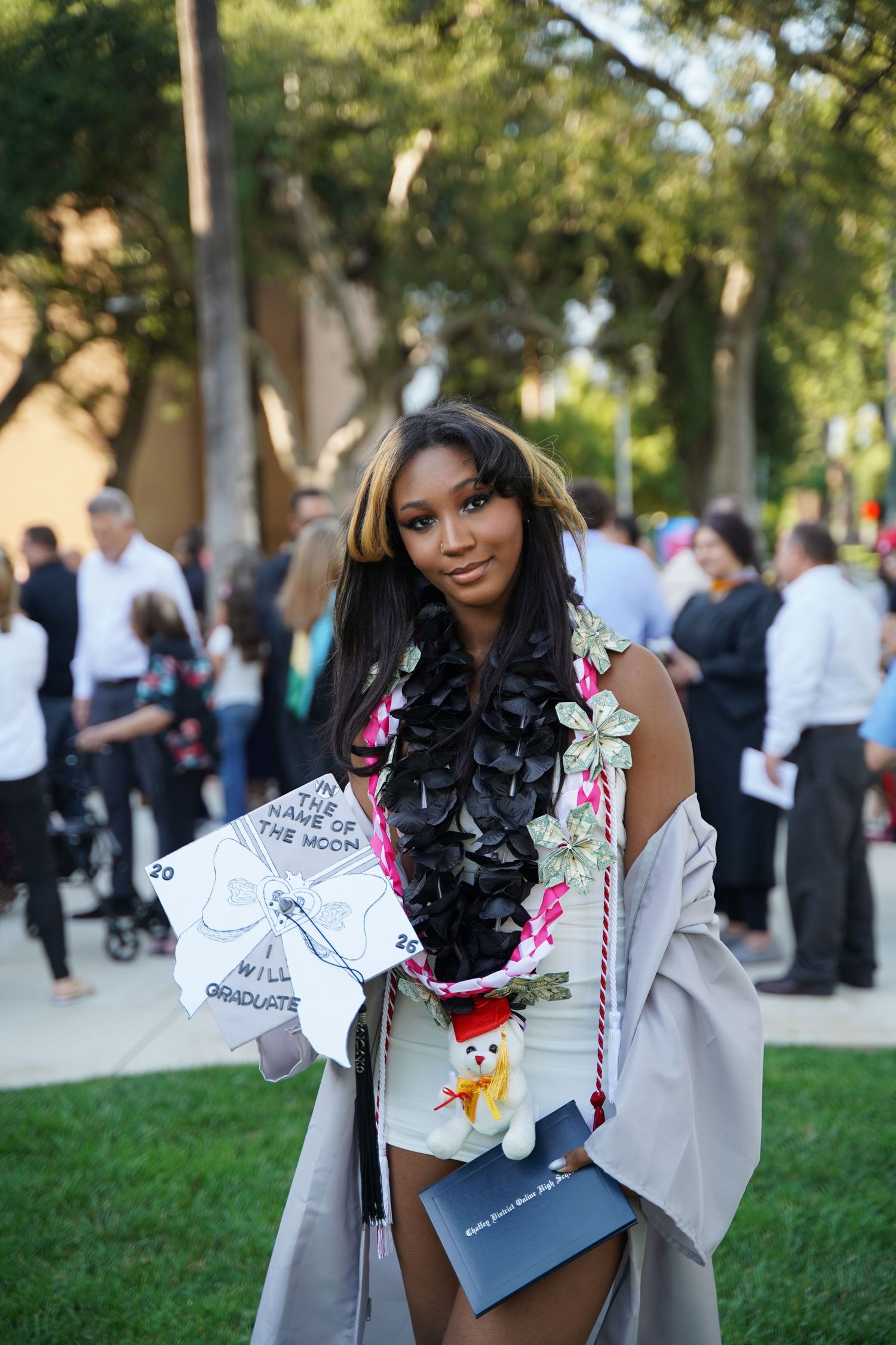 Young woman in graduation cap and gown smiles outdoors, decorated with leis and a stuffed animal at commencement ceremony.