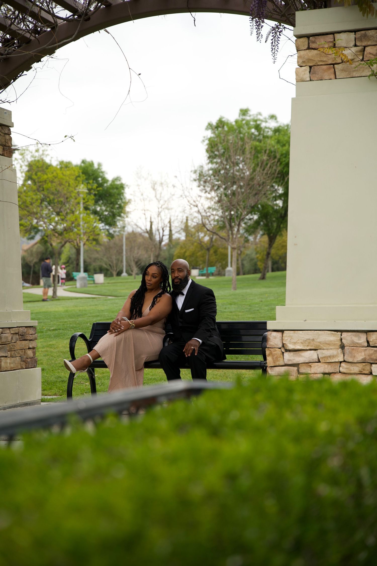 Elegant couple in formal attire sitting on a park bench under a stone pergola surrounded by lush green trees.